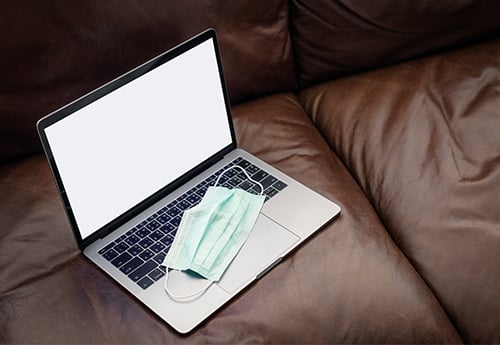 an open laptop on a leather couch with a surgical mask laying across the keyboard